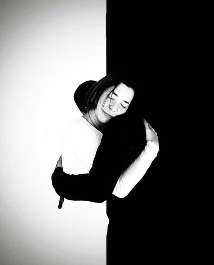 a black and white photograph of a couple embracing in front of a split background, half black and half white. the woman on the left side of the frame is wearing a white shirt and is hugging the man on the right side who is dressed in black. the man is holding the woman in his arms as they share a tender and affectionate moment. the image is full of contrast, creating a striking visual effect. the dark figure of the man against the white background and the bright face of the woman against the black background further emphasize the sense of unity and togetherness between them.