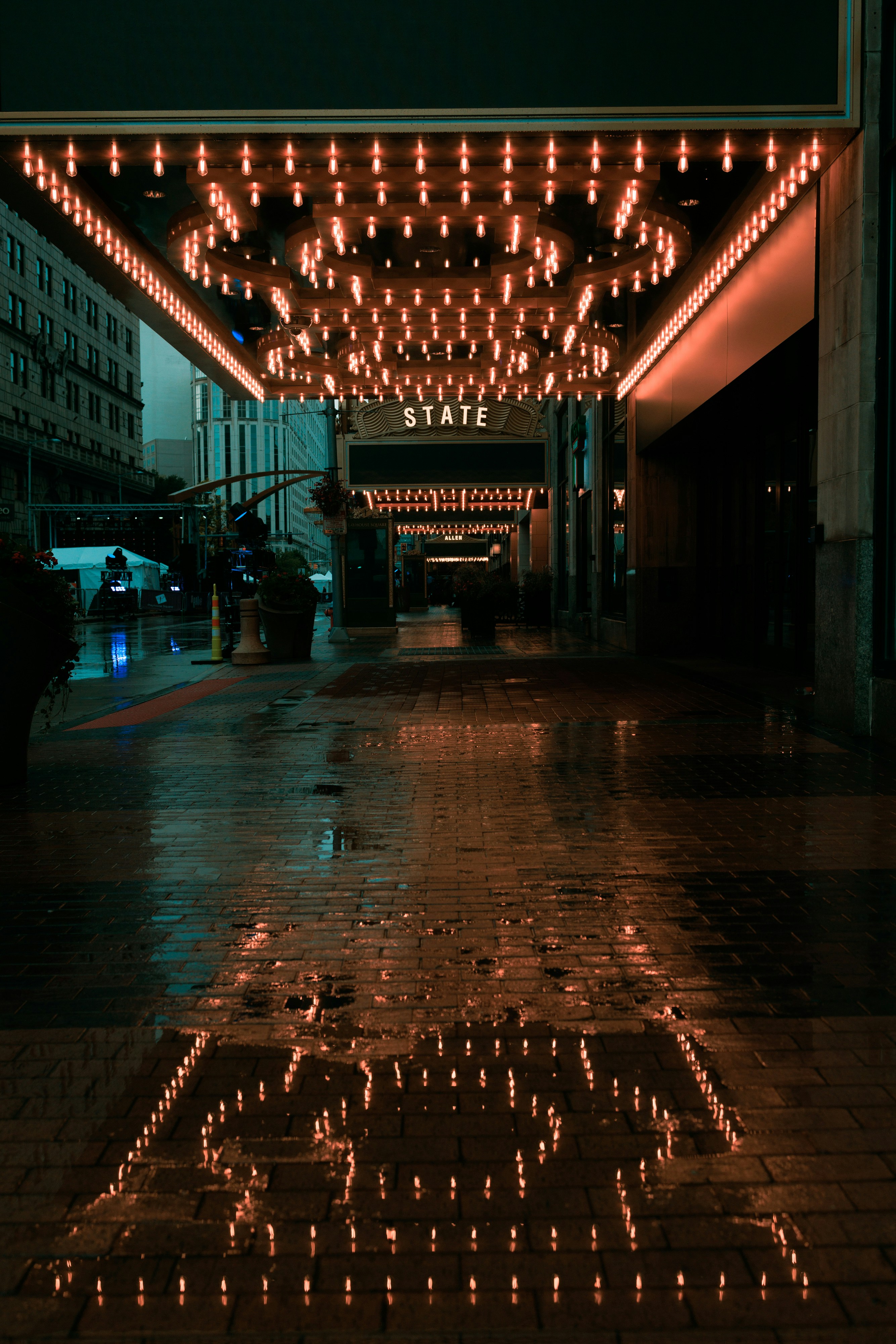 a city street at night after a rain. a building with a sign reading "state" is in the background, brightly lit with strings of lights under the awning. the wet sidewalk reflects the lights, creating a warm glow against the cool tones of the surrounding buildings and street. a puddle in the foreground reflects the lights most prominently, adding a sense of depth to the image.