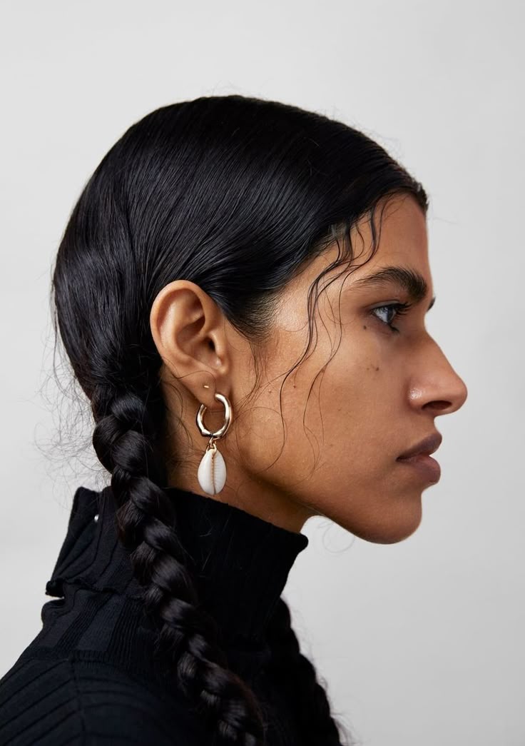 the image shows a profile of a woman wearing a gold hoop earring with a cowrie shell charm. she has dark hair styled in a braid and is wearing a black turtleneck sweater. the background is a plain gray wall.