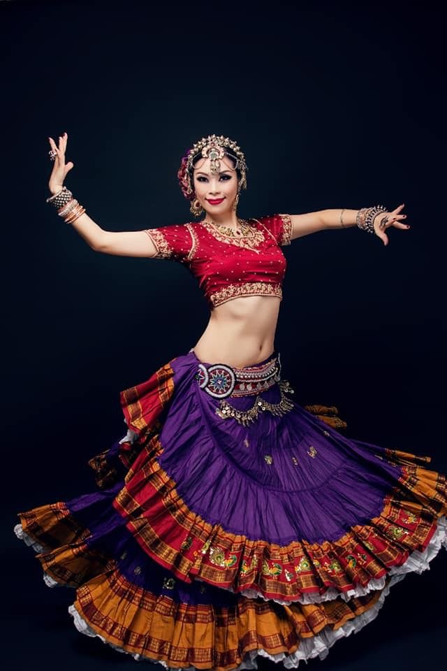 a young woman in a beautiful red and purple indian costume is dancing. she is wearing a traditional red top with gold embroidery and a flowing purple skirt with gold and red trim. her hair is adorned with jewelry, and she is wearing many other pieces of jewelry, including bangles on her wrists and rings on her fingers. she is posing dramatically with her arms outstretched, and she looks very elegant and graceful. the black background highlights the colors of her costume and her beauty.