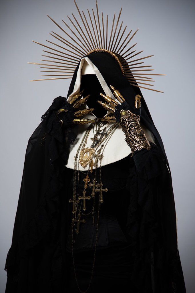 a person dressed as a nun, wearing a black robe and a gold halo, with a dark veil over their face, holding their hands up in front of them, covered in golden jewelry, with a variety of crosses hanging from their neck. the background is a plain grey color, and the overall vibe of the image is gothic and spooky.
