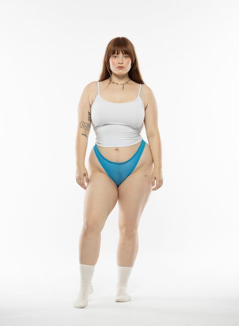 a plussize woman stands on a white background, wearing a white top, blue underwear, and socks. she has brunette hair with bangs and is wearing a necklace. she has tattoos on her arms and is standing indoors.