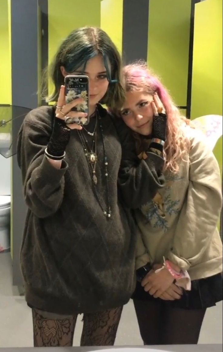 a selfie of two teenage girls posing in a bathroom, one of the girls has green hair and is wearing a brown sweater, black fishnet stockings, and a pearl necklace. the other girl is wearing a white sweater and has pink hair. both girls are wearing black gloves. they appear to be best friends.