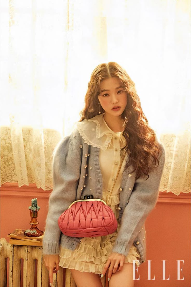 a woman with brown, curly hair is posing for a portrait. she is wearing a blue cardigan with rhinestones, a ruffled blouse, and earrings. she is also wearing a pink, quilted purse on a strap. the background includes vintage curtains, a lit candle, and the elle magazine logo. she is looking at the camera.