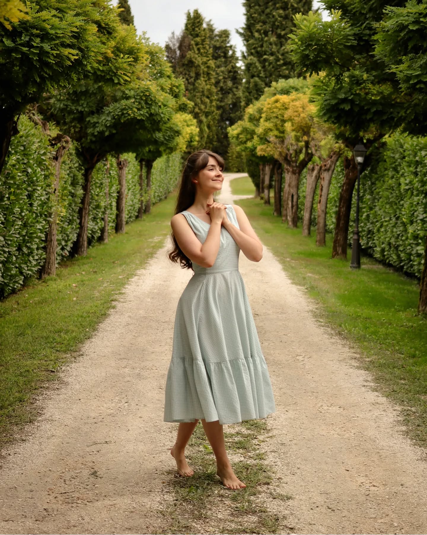 a young woman in a light blue linen midi dress walks barefoot down a gravel path lined with lush green trees.  she is looking up with a peaceful and joyful expression, her hands clasped together near her chest. the scene is idyllic and serene, suggesting a feeling of calm and happiness. the natural light creates a warm and inviting atmosphere.