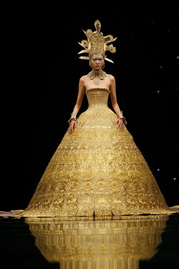 a model is standing on a reflective stage, wearing an elaborate golden gown and headdress. the gown has a large, full skirt and ornate details. the headdress is tall and intricate. the background is dark with faint stars. the image captures a fashion show atmosphere with theatrical elements.