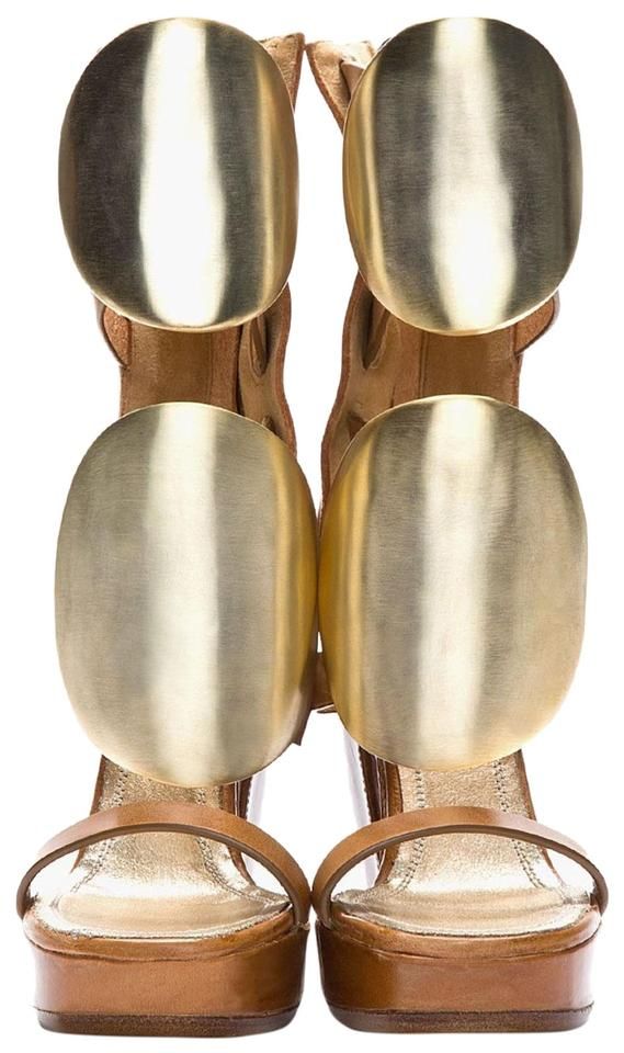 a closeup shot of a pair of women's platform sandals. the sandals are light brown leather and feature large, ovalshaped gold metallic accents on the straps. the heels are chunky and the sandals have a unique, statementmaking design. the background is plain white.