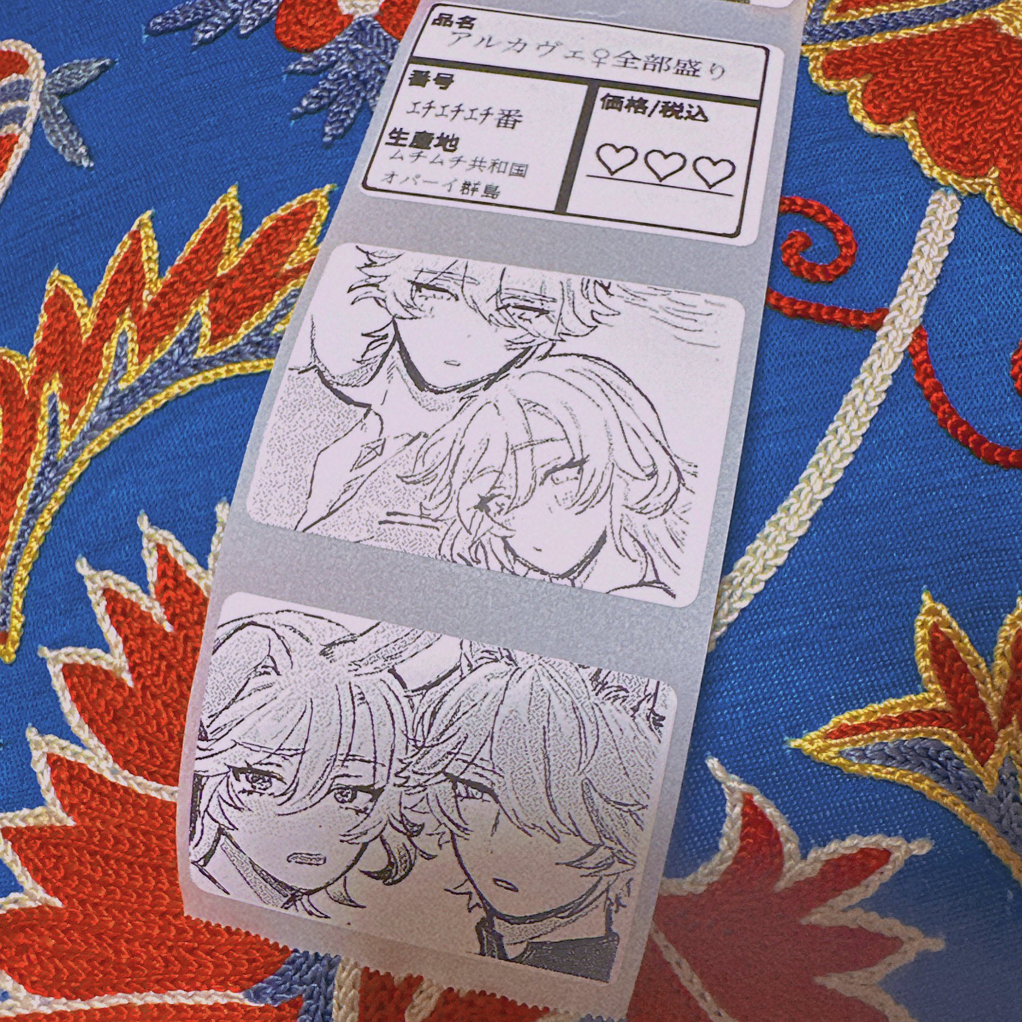 a closeup shot shows a price tag with anime artwork affixed to a piece of fabric with red, gold, and blue floral embroidery. the price tag includes text in japanese and two panels with cartoon illustrations of anime characters. the background fabric is blue with detailed stitched patterns of flowers and leaves.