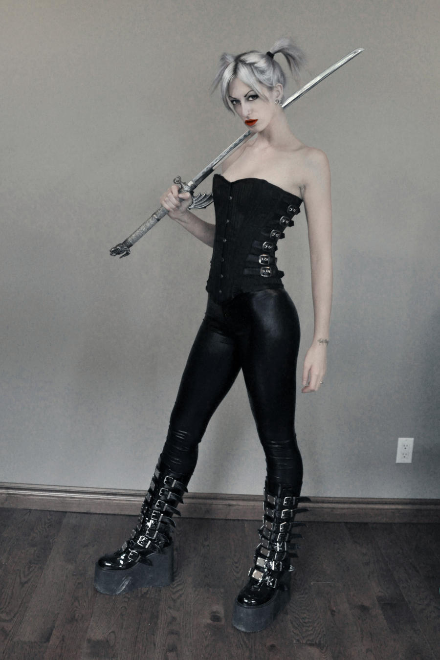 a fulllength shot of a young woman with pale skin and white hair styled in pigtails. she's dressed in a dark gothic style outfit consisting of a black corset, black leggings, and tall, black platform boots with multiple buckles. she holds a silver sword across her shoulders, striking a fierce and confident pose.  the background is a simple, neutral grey. the overall aesthetic is dark, edgy, and dramatic.
