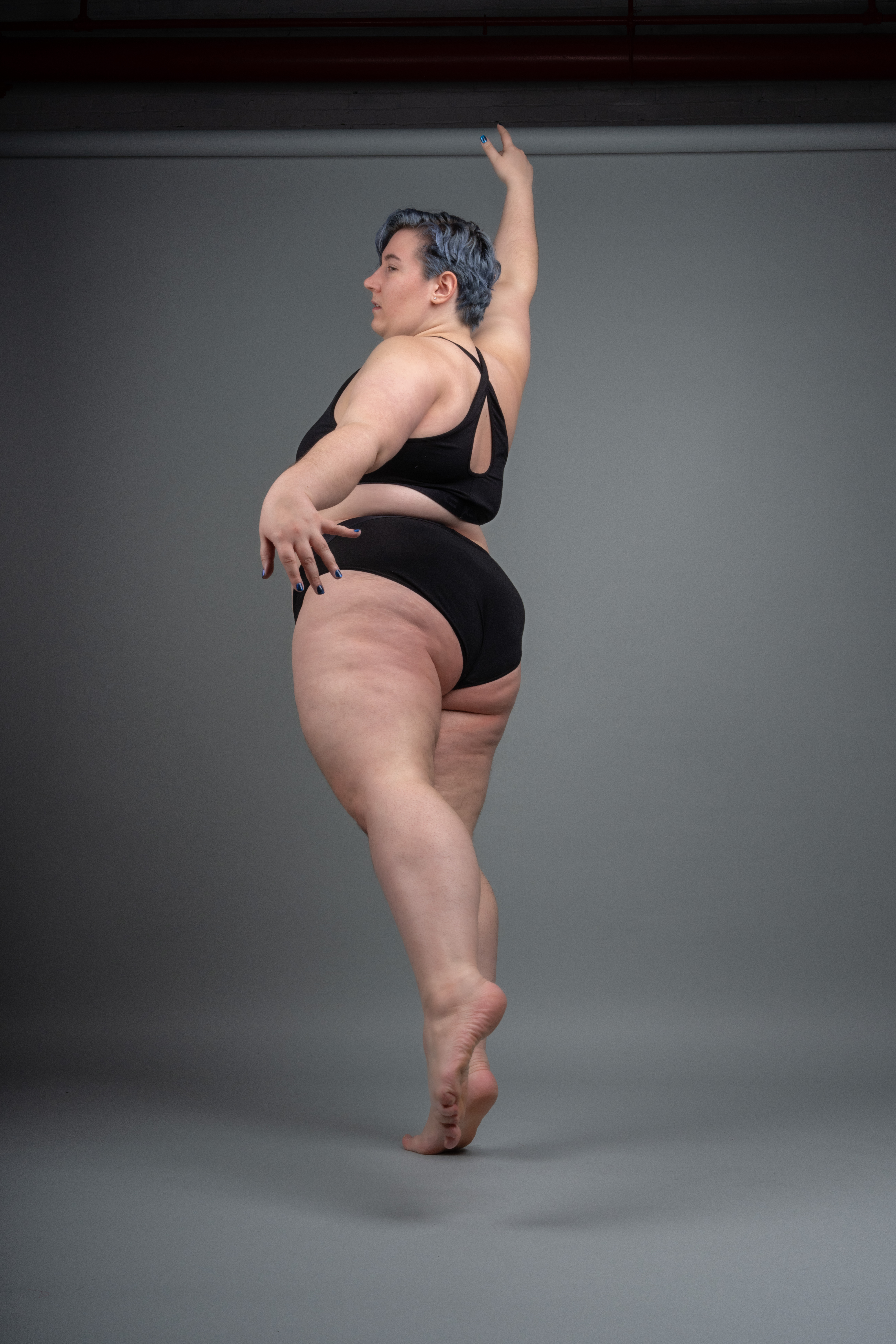 a plussize woman with blue hair stands in a dance pose against a gray background. she is wearing black underwear and has one arm raised. her body is facing to the side, showing her full figure. the photo appears to be a studio shot intended to promote body positivity.