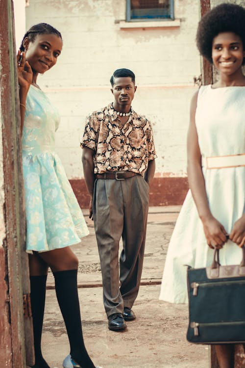 the image shows three young adults, two women and one man, standing outdoors. the women are wearing vintageinspired dresses – one light blue, one offwhite. the man is wearing a printed shirt and grey trousers. all three are of african descent. the scene is set against the backdrop of a building with colonial architecture.  one woman is partially hidden behind a doorway. the image has a retro or vintage feel.