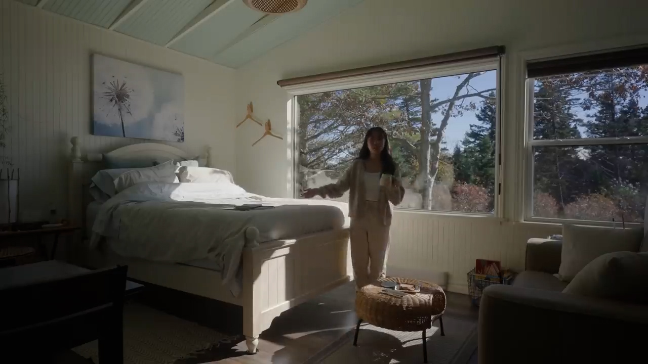 a woman is standing in a brightly lit bedroom with a mug in her hand.  the room has a large bed with white bedding and a wooden frame. there are large windows that look out onto trees. a woven coffee table sits in front of the windows with a book and other items on it. to the right is a couch with white pillows. the overall feel of the image is bright, airy, and peaceful.