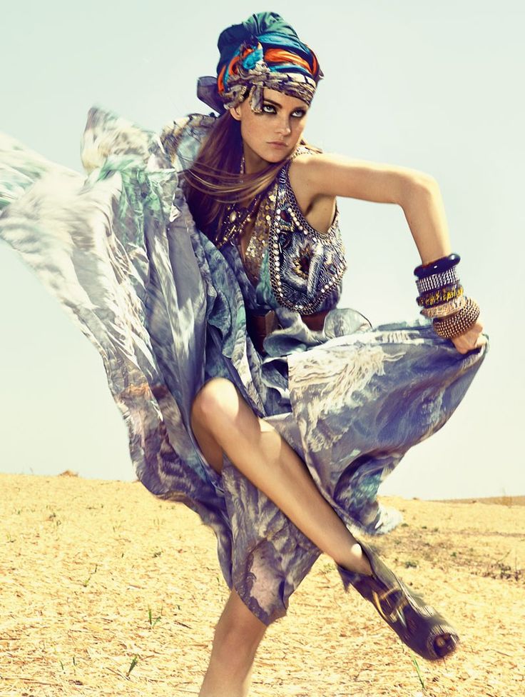 a fashion model in a colorful dress and head wrap stands in a desert environment. the model is looking fierce and stylish.  the dress is long and flowing, and the model is wearing bangles and bracelets on both wrists.  the model's hair is long and brown.
