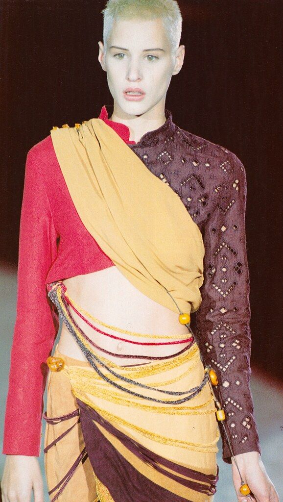 a runway model is shown from the waist up, wearing an avantgarde highfashion outfit that showcases an asymmetrical and colorblocked design. the model, who has short, lightcolored hair and an androgynous look, is wearing a top that is half red with a full sleeve and half brown with a decorative perforated pattern and a full sleeve. a goldcolored fabric is draped over the upper body, creating a shawl effect. the garment exposes the midriff. the skirt part of the outfit is also asymmetrical and colorblocked, with gold and brown panels, accented by strands of amber beads that hang around the waist and hip. the photograph captures a moment from a fashion show, highlighting the unique and artistic qualities of the clothing.