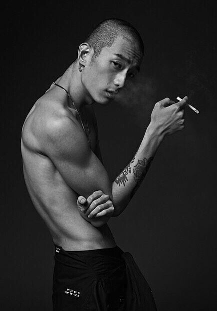 a shirtless man with a tattoo on his arm poses for the camera while holding a cigarette in his right hand. he has his arm bent at the elbow and is resting his hand on his bicep, holding a lit cigarette in the other hand. he is looking at the camera. the image is in black and white.