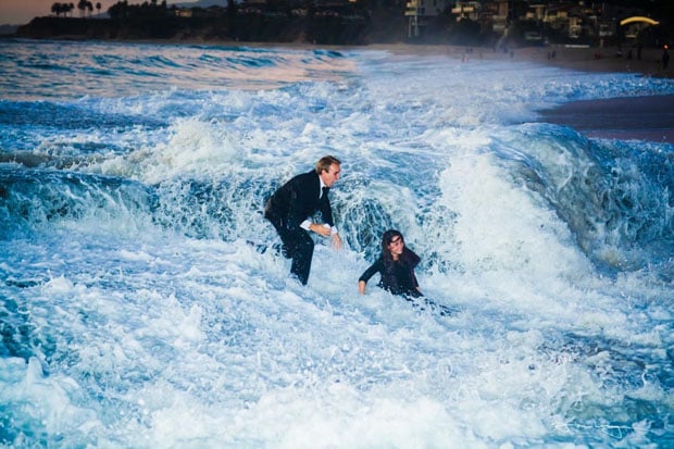 a man and a woman, both wearing business suits, are caught in crashing ocean waves. the scene takes place at a beach with the remnants of daylight illuminating the sky. the overall palette is cool toned, emphasizing the blue of the water and the white of the crashing waves.  the man is attempting to help the woman stay afloat, creating a sense of struggle against the powerful waves. this juxtaposition of formal attire and a chaotic natural setting suggests a narrative of playfulness, challenge, or perhaps a commentary on the unpredictable nature of the business world.