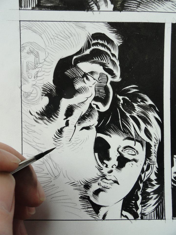 a comic book artist is inking a closeup panel of a female character's face with a brush. the character is drawn in a realistic style, and the inking is very detailed. the artist is working on a piece of paper, and the panel is clearly visible in the image. the artist's hand is holding the brush, and the tip of the brush can be seen in the image. the artist is wearing a blue shirt.  the character has an expression of surprise or awe.  the artist is creating a beautiful and complex comic book illustration. the process is being done with a great deal of care and skill.