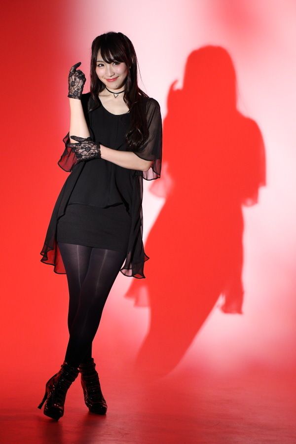 a woman stands against a red background, casting a shadow behind her. she's wearing a black dress, black tights, and black highheeled boots. lace gloves adorn her hands, and a choker necklace circles her neck.