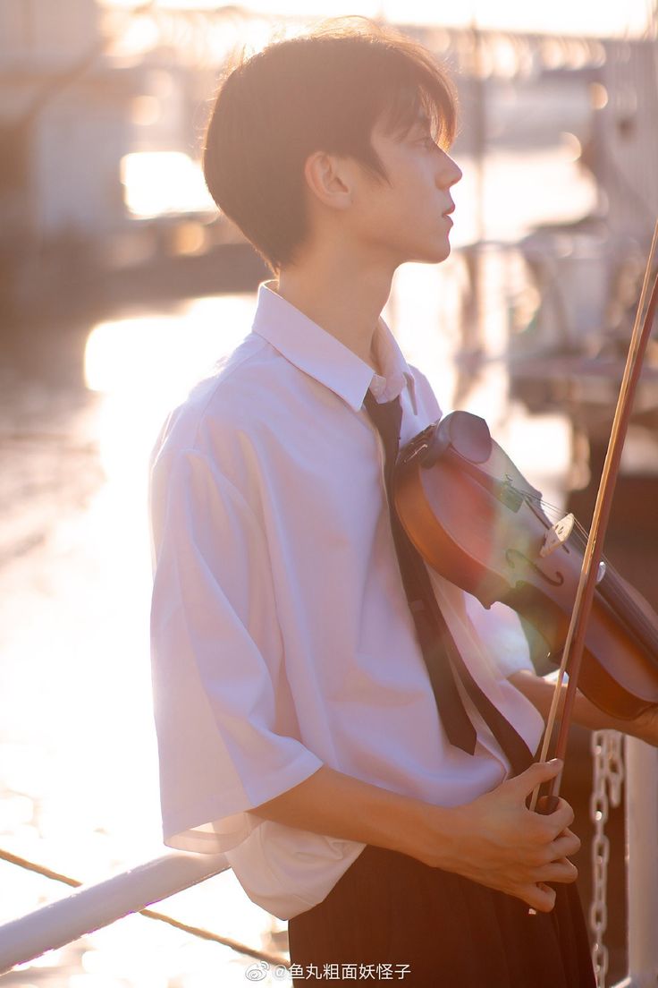 a young asian man wearing a white shirt and tie stands on a pier by a body of water, holding a violin. he is backlit by the setting sun, creating a warm and serene atmosphere.