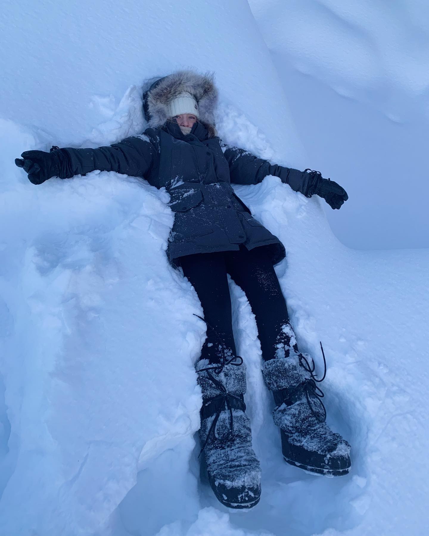 a girl is lying in the snow, making a snow angel. she is wearing a dark gray parka, a white hat under a fur hood, dark gloves, and dark snow boots. she is smiling and looks happy.  the snow is deep and fluffy, and she has created a distinct snow angel shape around her body. the scene is peaceful and wintery.