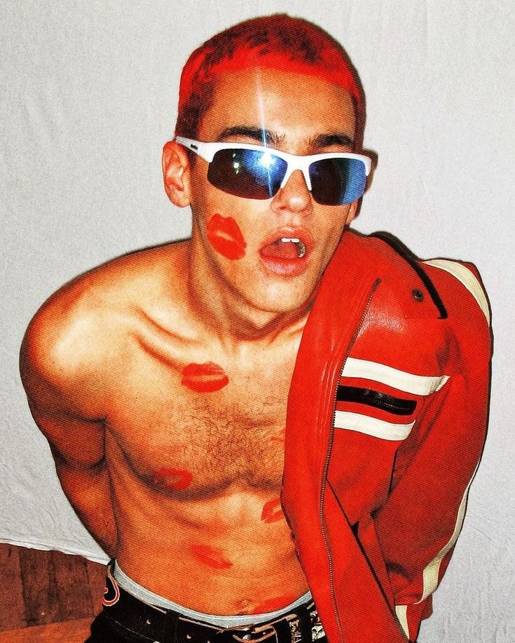 a young man with bright red hair and a red leather jacket poses with his shirt off. he is wearing sunglasses with a white frame and red lip prints on his chest. he is looking towards the camera.