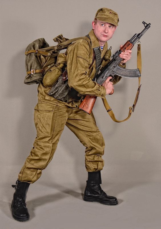 a soldier wearing a military uniform and backpack is holding an ak47 assault rifle. he is looking to his right side. the soldier is standing on a light grey background.