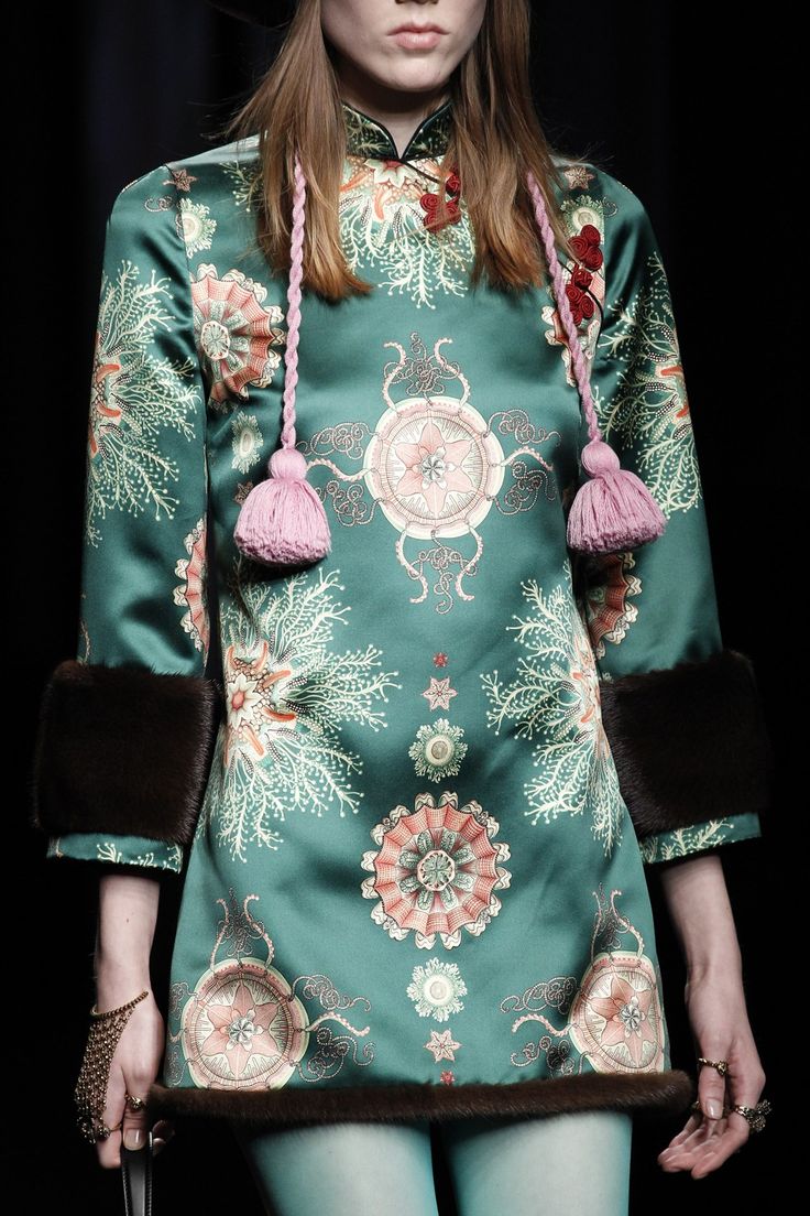 loseup shot of a model on a fashion runway wearing a teal silk dress with a detailed floral print. the dress has a mandarin collar, long sleeves with fur cuffs, and pink tassels hanging from the collar. the model is wearing teal tights and jewelry, including bracelets and rings. the overall style is elegant and luxurious, showcasing the intricate detail of the gucci design. the background is dark and blurred, focusing attention on the dress and model.
