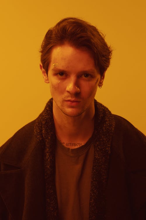 the image is a closeup portrait of a young man with dark brown hair. he is looking directly at the camera with a serious, almost intense expression.  he has a small bandage or scar on his left cheekbone and a small tattoo visible on his neck. he's wearing a darkcolored jacket or cardigan over a muted brown tshirt. the lighting is warm, ambertoned, creating a moody and dramatic atmosphere. the background is a solid, pale yellow. the overall impression is one of seriousness and perhaps a hint of vulnerability.