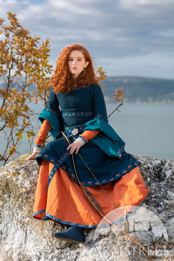 a young woman with curly red hair dressed in a medievalstyle costume sits on a rock outdoors, holding a bow. she wears a blue dress with orange sleeves and a long orange skirt underneath. the background includes a lake, trees with autumn foliage, and a cloudy sky.