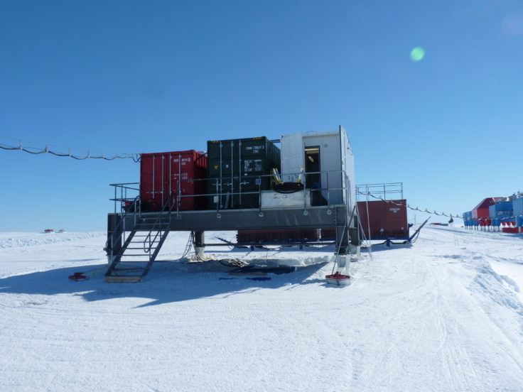 an image of a research station in antarctica with multiple containers and buildings, all built on platforms to keep them above the snow. the sky is clear and blue, and the ground is covered in snow.