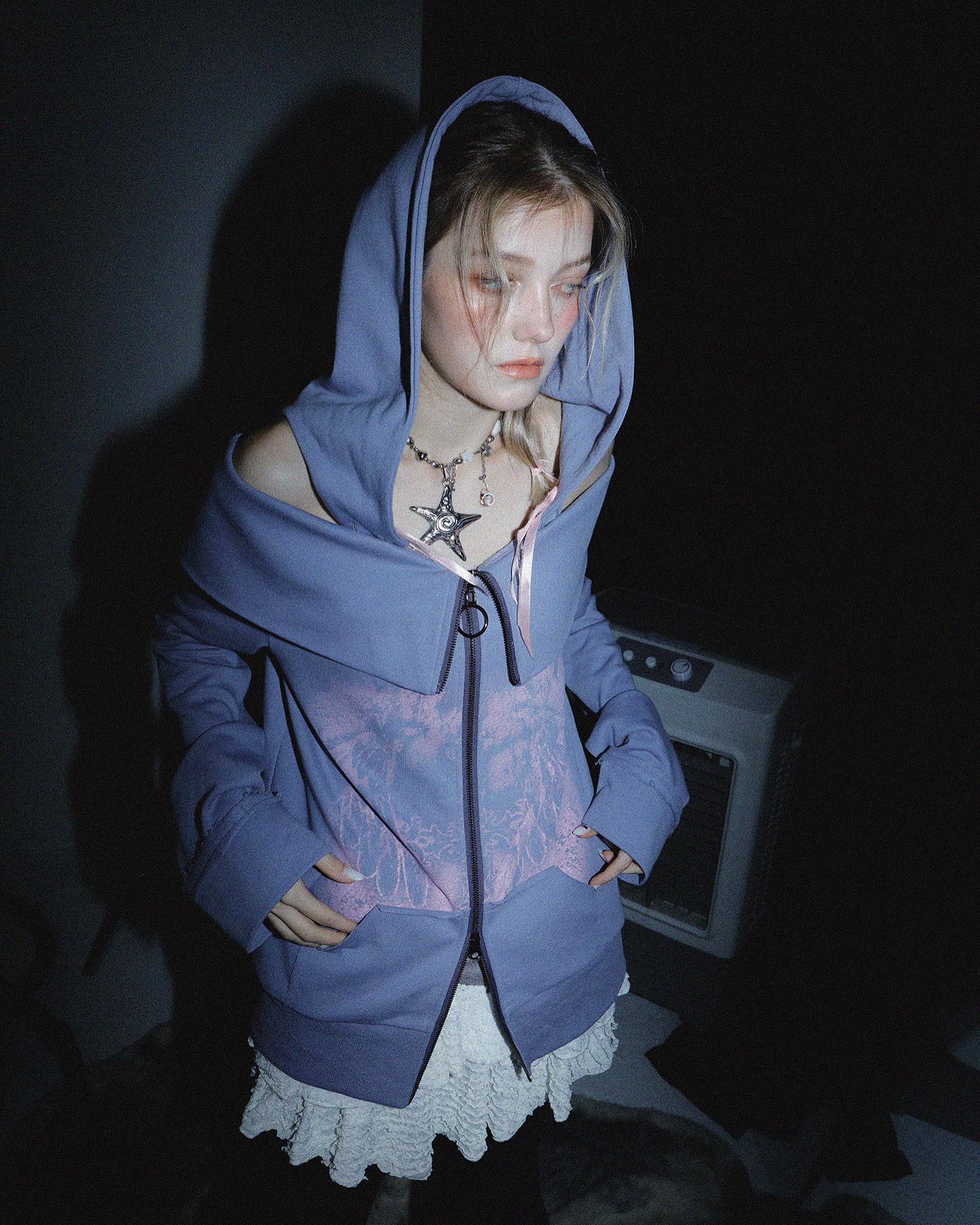 a young woman with blonde hair is captured in a closeup, eyelevel indoor shot. she's wearing a stylish, offtheshoulder lavender hoodie with a subtle pastel print, paired with a white lace skirt. the hoodie features a zipper, a hood, and pockets, and the overall style is a blend of grunge and alternative fashion. her accessories include a starshaped necklace and subtle makeup. the background is dark and moody, enhancing the atmospheric feel of the photograph. the image has the aesthetic of a vintage film photograph.