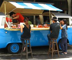 a brightly colored food truck is parked on a city street. the truck is painted blue and has a striped awning. there is a man and a woman standing in front of the truck. the man is talking to a group of customers while the woman is serving them. the truck is filled with food and drinks, and it is open for business.