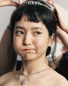 a young woman with a straightcut black bob and bangs is the subject of a closeup vogue photoshoot. she is wearing a delicate silver necklace with a teardrop pendant, and a headband adorned with the word "vogue" in sparkling letters. her expression is slightly pouty, with her lips pursed.  two hands with redpainted fingernails are shown adjusting the headband atop her head. the background is blurred, suggesting a studio setting with professional lighting illuminating her face.