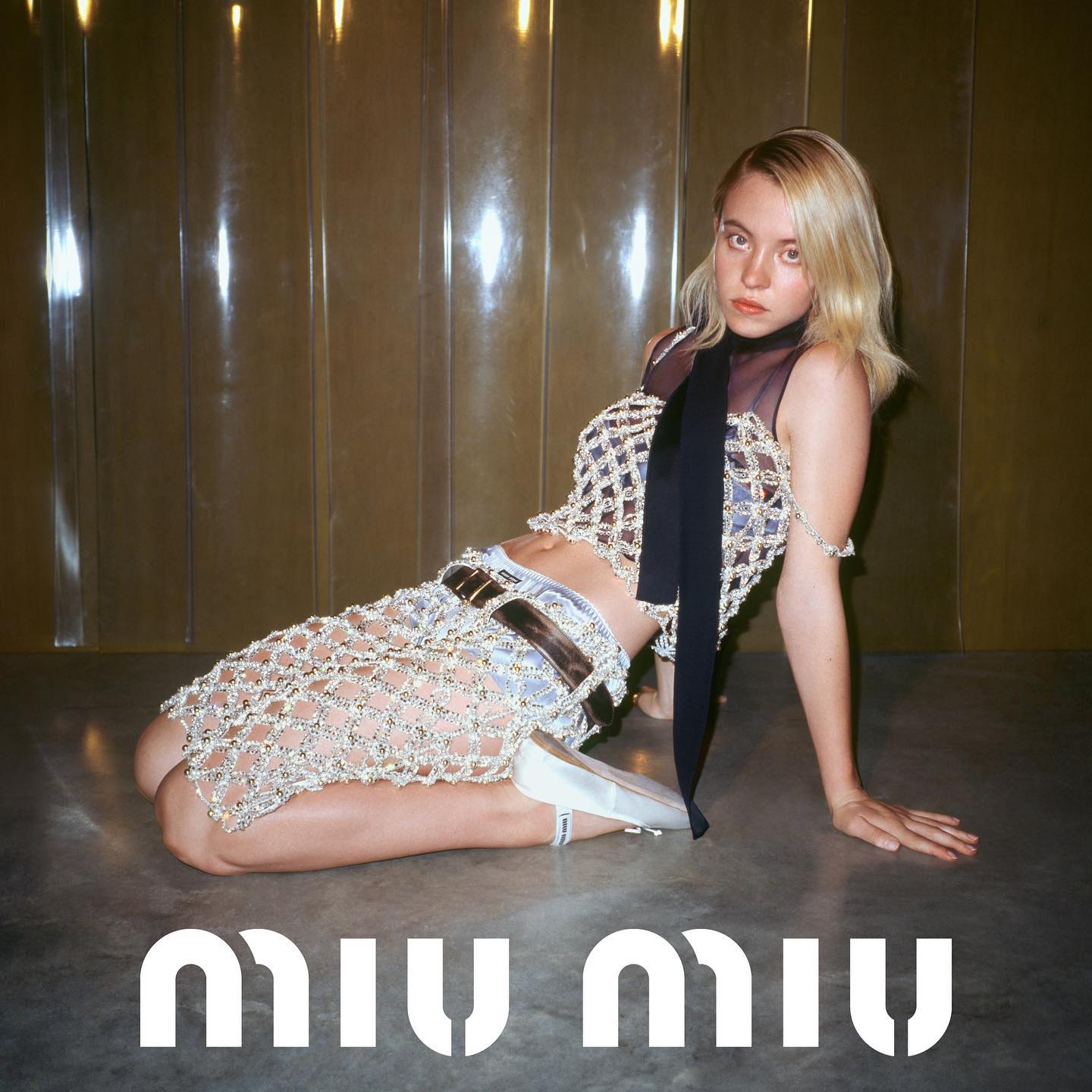 the image is a fashion advertisement for miu miu. it features a young blonde woman posing on a floor with a goldtoned wall in the background. she's wearing a stylish outfit consisting of a beaded, netlike skirt layered over denim shorts, a matching cropped beaded top, and a dark scarf. the model wears lightcolored flat shoes and is posed in a relaxed yet elegant way.  the overall aesthetic is high fashion and contemporary.  the miu miu logo is prominently displayed at the bottom of the image.