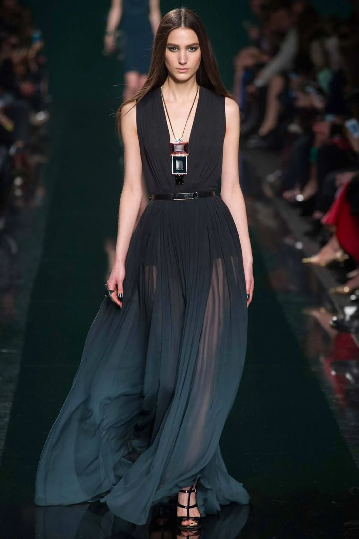 a fashion model walks the runway in a long black and green dress with a sheer overlay. she is wearing a silver necklace with a black pendant and high heeled black sandals. the model is posing for the camera with a confident expression. the background is blurred and shows a runway with a dark green background.