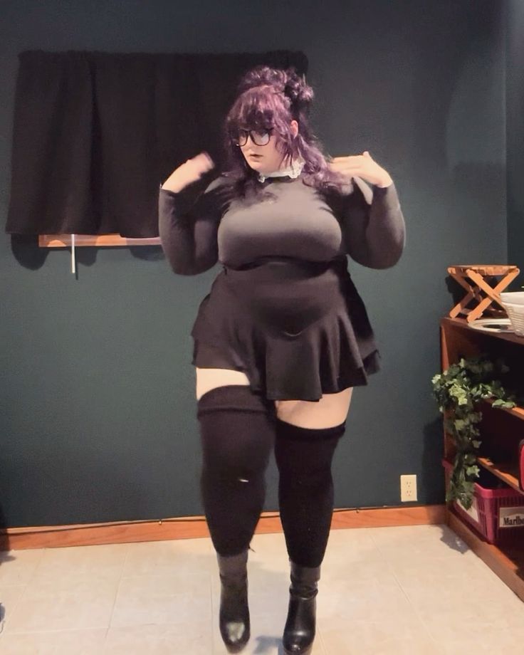 a woman with purple hair and glasses, wearing a black dress, thighhigh socks, and boots, is standing indoors.