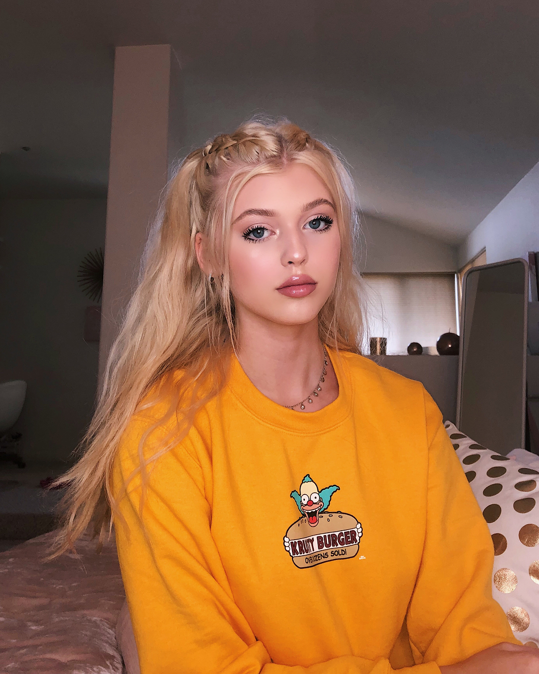 a portrait of a young woman with blonde hair styled in pigtails and braids wearing a yellow krusty burger sweatshirt. she has blue eyes and is looking at the camera.