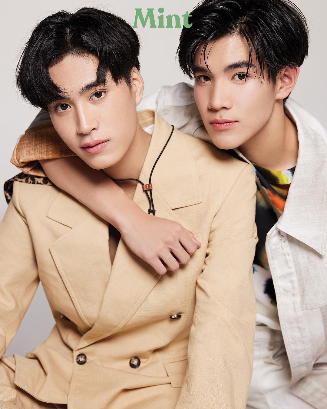 two young asian men pose together for a fashion photoshoot. one man, wearing a beige doublebreasted suit, sits slightly forward with a relaxed posture. his left arm is draped over the shoulders of the other man, who stands behind him. the second man, dressed in a white shirt and a patterned overshirt, leans slightly forward and rests his right hand on the first man's arm. both men smile gently at the camera, exuding a sense of camaraderie and effortless style. the backdrop is a clean white, allowing the focus to remain on the models and their clothing.