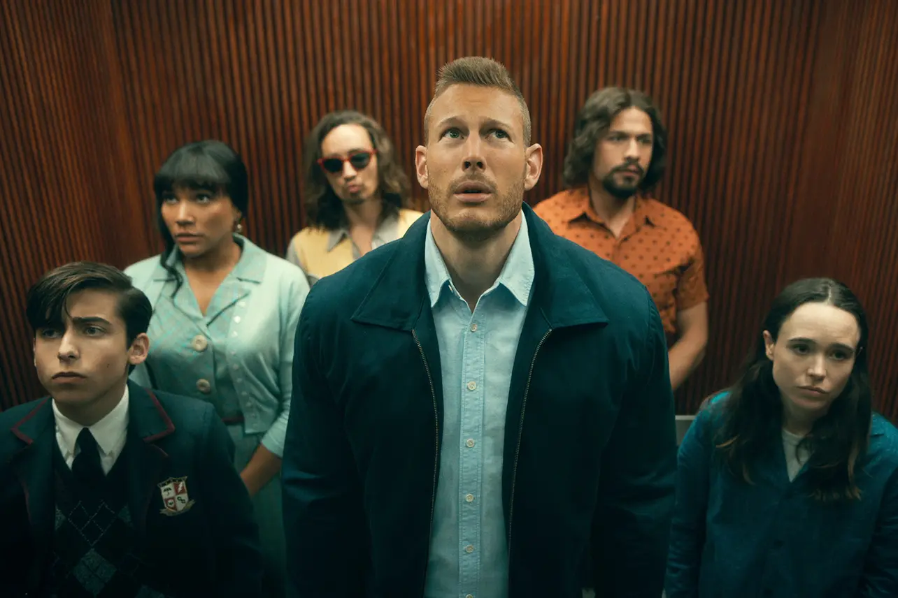 a lowangle shot of the main cast of the tv show the umbrella academy standing in an elevator and looking up.  the elevator has dark woodpaneled walls and a dark ceiling. the lighting is artificial and yellowish.