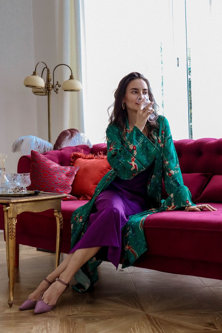 a stylish woman is sitting on a luxurious red velvet sofa in a beautifully decorated room. she's wearing a vibrant green silk robe over a purple dress and is holding a wine glass, taking a sip. the overall ambiance is elegant and sophisticated, with the woman radiating confidence and poise. the room features a hardwood floor, elegant curtains, and a gold floor lamp. the woman's attire, posture and the setting all contribute to the image's overall luxurious feel.