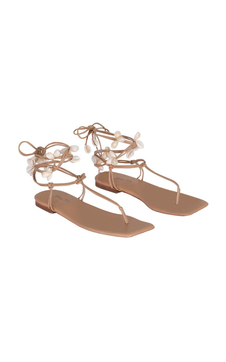 the image shows a pair of women's beige flat sandals. the sandals are embellished with small white shelllike decorations along the laceup straps. the straps wrap around the ankle and across the foot, securing with a tie closure. the sandals have a simple, minimalist design with a squaredoff toe.  they appear to be made of a smooth, possibly leatherlike material. the overall style is summery and casual, suitable for beach or warm weather wear.