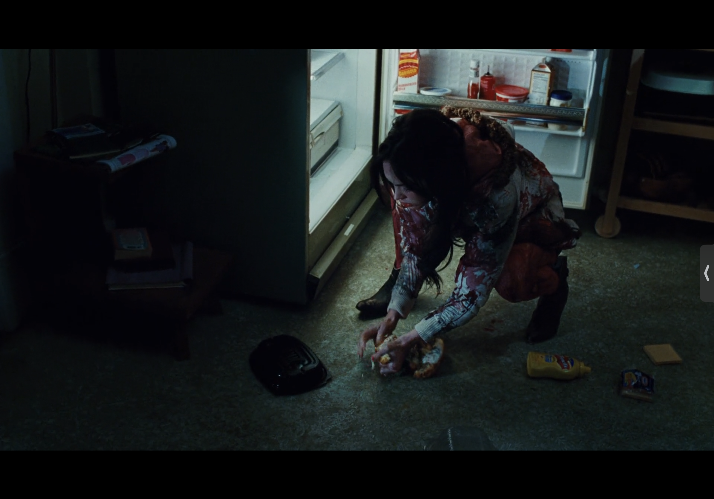 a woman with long, dark hair is kneeling on the floor of a kitchen. she is wearing a red and white bloody shirt and is eating a sandwich. she looks down at the sandwich and the floor, and the kitchen appears dark and scary. there is a refrigerator in the background and other items on the floor.