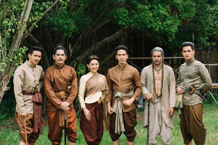 a group of 6 people, 5 men and 1 woman, are standing in a garden setting. the people are all wearing traditional thai clothing. the men are wearing longsleeved shirts and pants, while the woman is wearing a dress with a long scarf draped over her shoulders. the people are smiling and looking at the camera. there are trees in the background, and the ground is covered in grass. there is a wooden stick in the middle of the image and a sword in the right side.