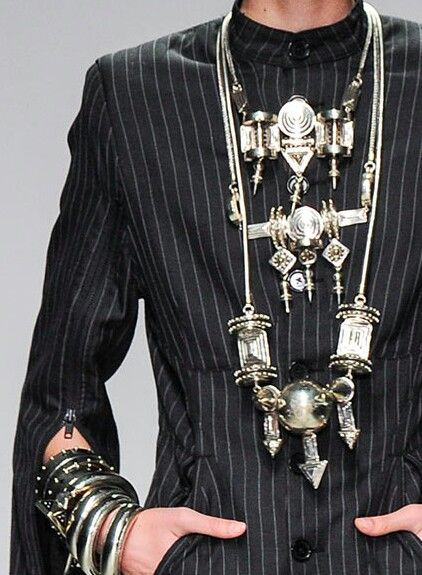 a person is wearing a black pinstripe suit with an elaborate multilayered necklace and multiple metal bracelets. the suit jacket has a zipper detail near the cuffs, and the person's hands are partially visible.