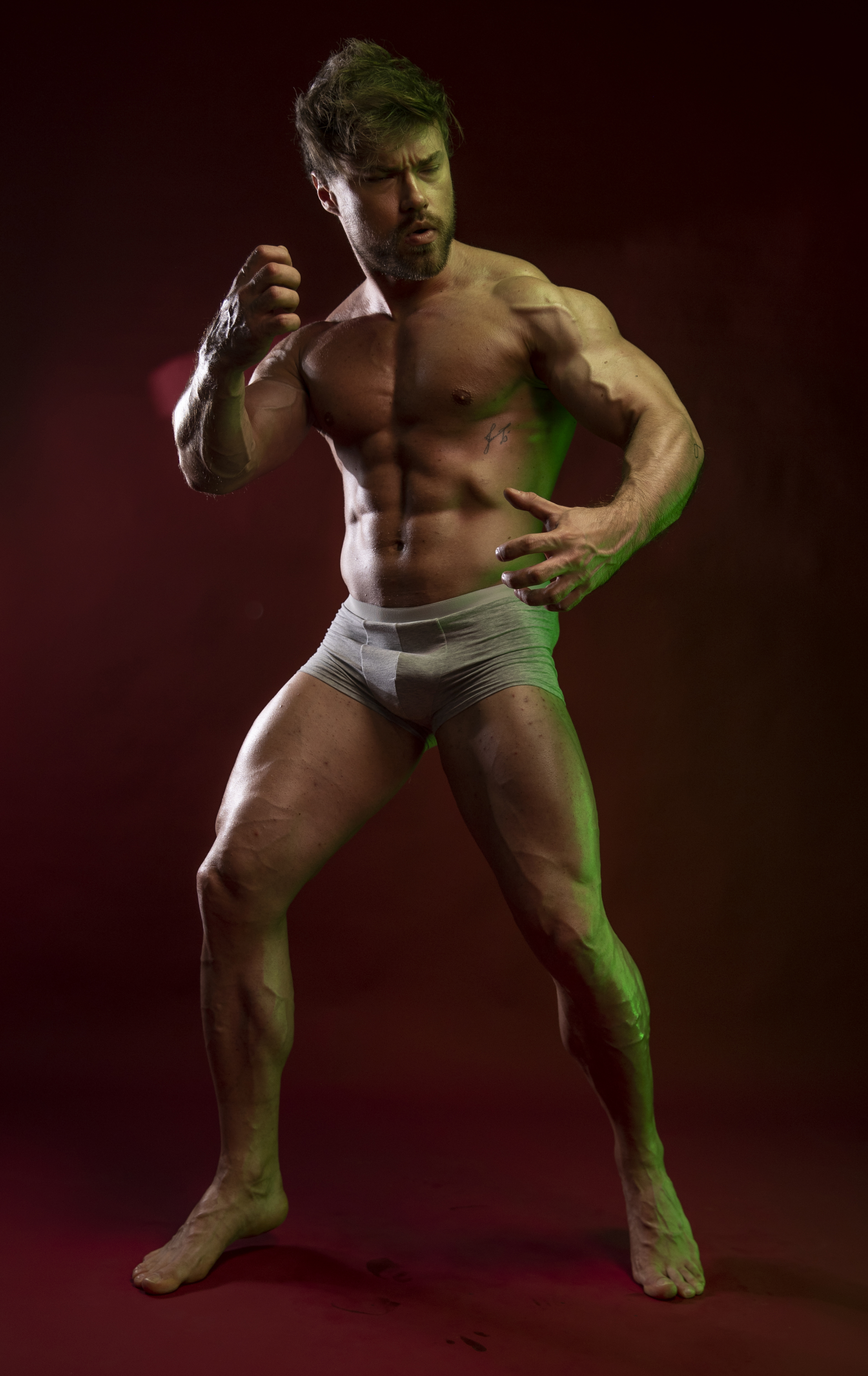 a muscular, shirtless man wearing underwear stands on a red background illuminated by green lighting. he is posing with a serious expression, fists clenched, showcasing his physique and defined abs. his bare feet are visible, and veins are prominent on his body.