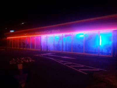 a night shot of an empty car wash building illuminated with neon pink and blue lights. the building is modern and sleek, with large glass windows. the neon lights reflect off the wet pavement in front of the building.