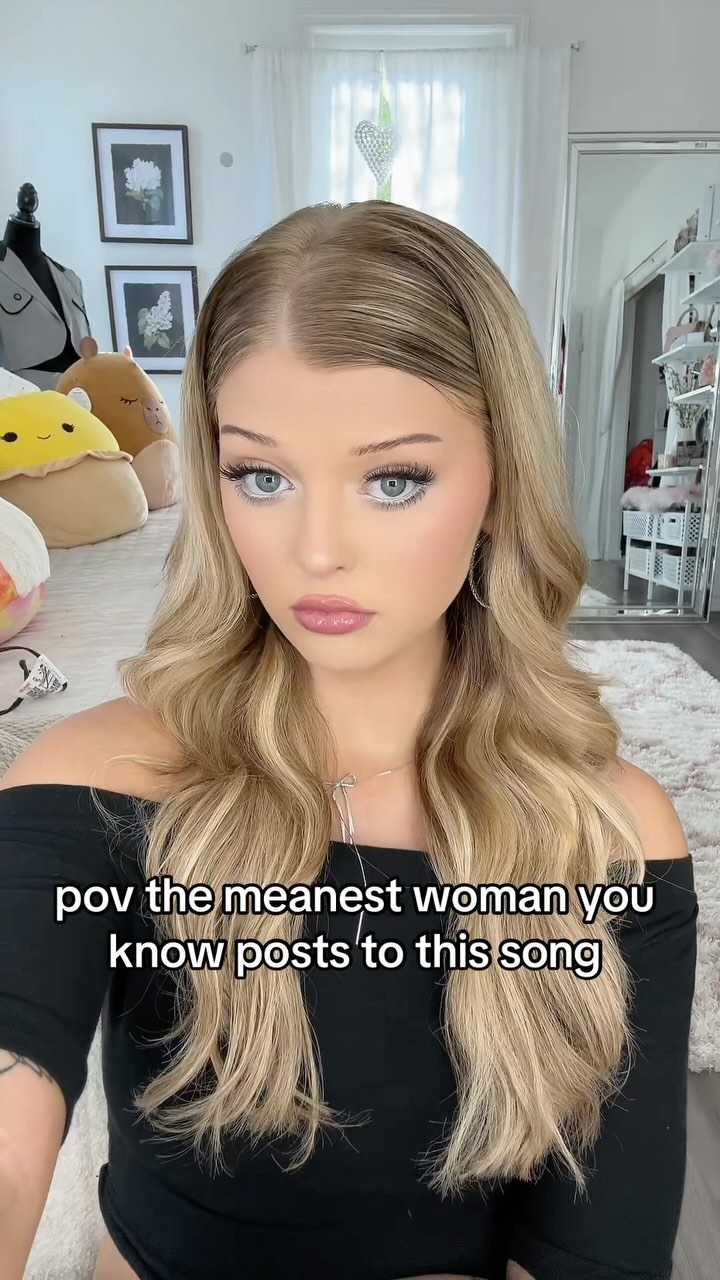 a selfie of a blonde woman wearing a black offtheshoulder top and jewelry. she is in a room with a couch and squishmallows behind her. the text "pov the meanest woman you know posts to this song" is overlayed on the image.