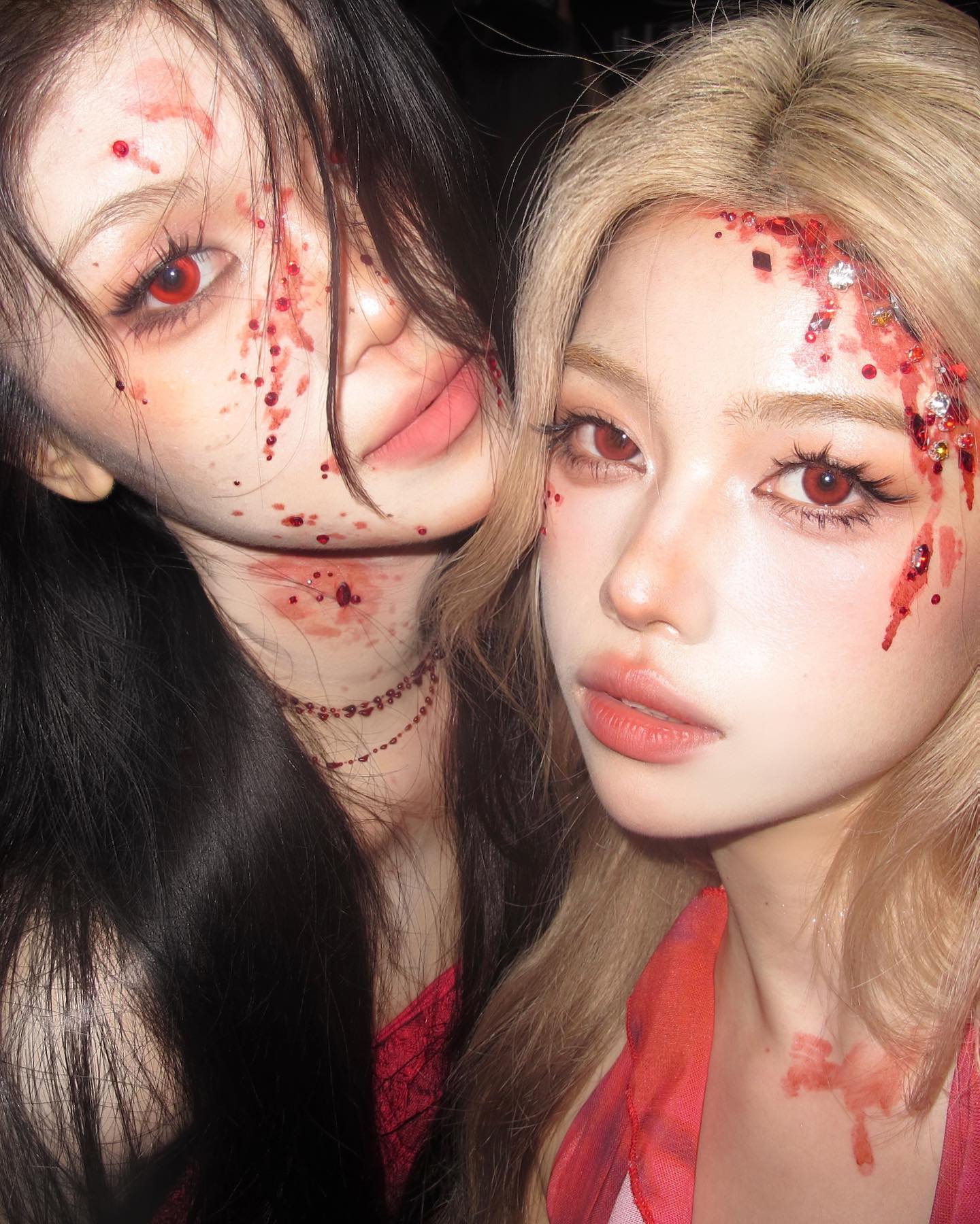 two young women pose for a selfie in their halloween costumes.  they are wearing glamorous makeup with fake blood and crystals. the woman on the left has long, black hair, while the woman on the right has long, blonde hair.  the overall aesthetic of the photo is glamorous and spooky.