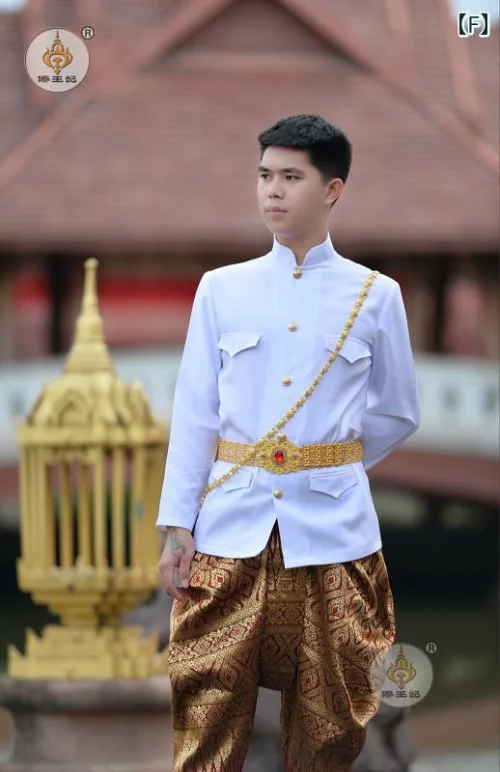 a thai man wearing a traditional white outfit with gold accents stands in front of a gold structure, smiling at the camera. he is wearing a white longsleeved shirt with gold buttons and a gold sash, and gold pants with a pattern.