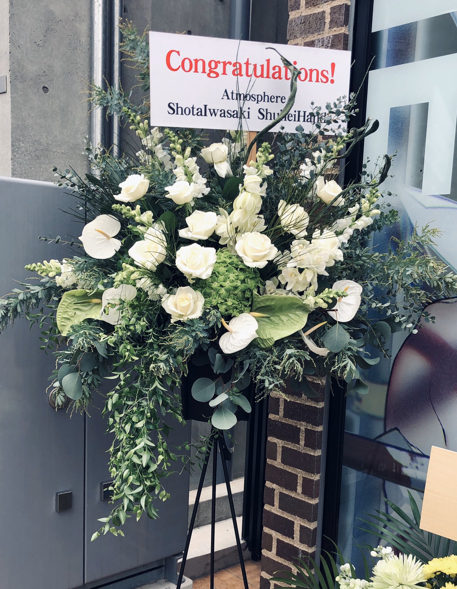 a large floral arrangement stands in front of a brick wall. the arrangement features white roses, lilies, and greenery, arranged in a tall, rectangular vase. a sign reading "congratulations!" is prominently displayed at the top, followed by the names "atmosphere," "shota iwasaki," and "shuhei." the scene suggests a celebration, possibly a grand opening or similar event.